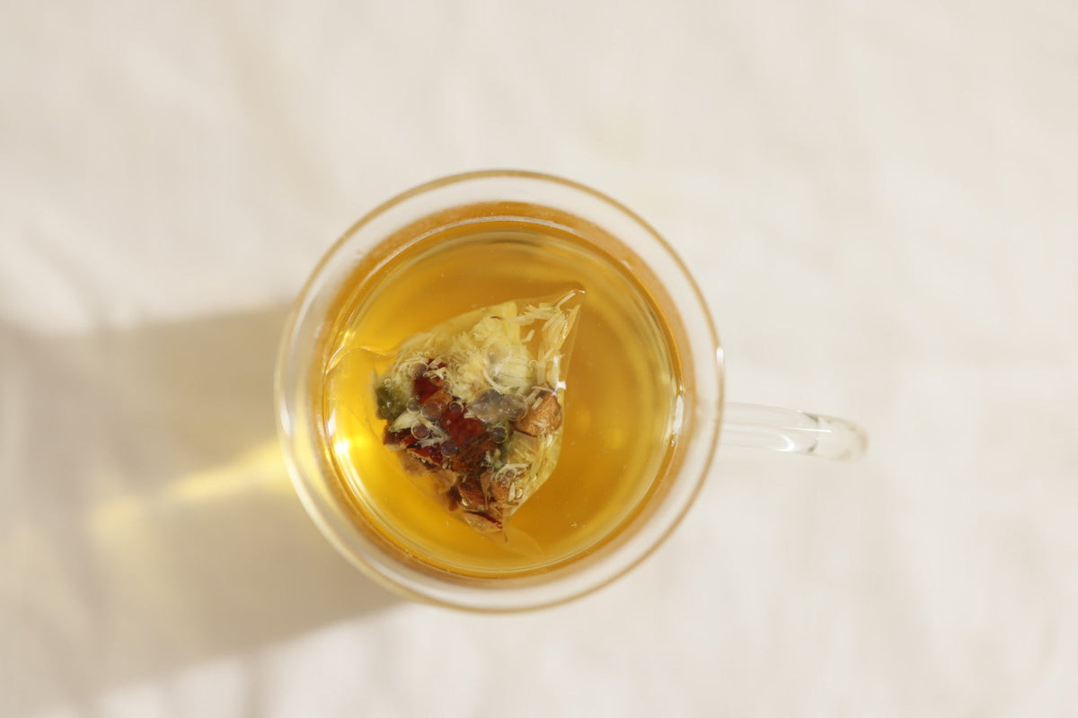 An oversized sachet of qìsane’s chrysanthemum & goji steeping in golden tea in a clear glass cup.