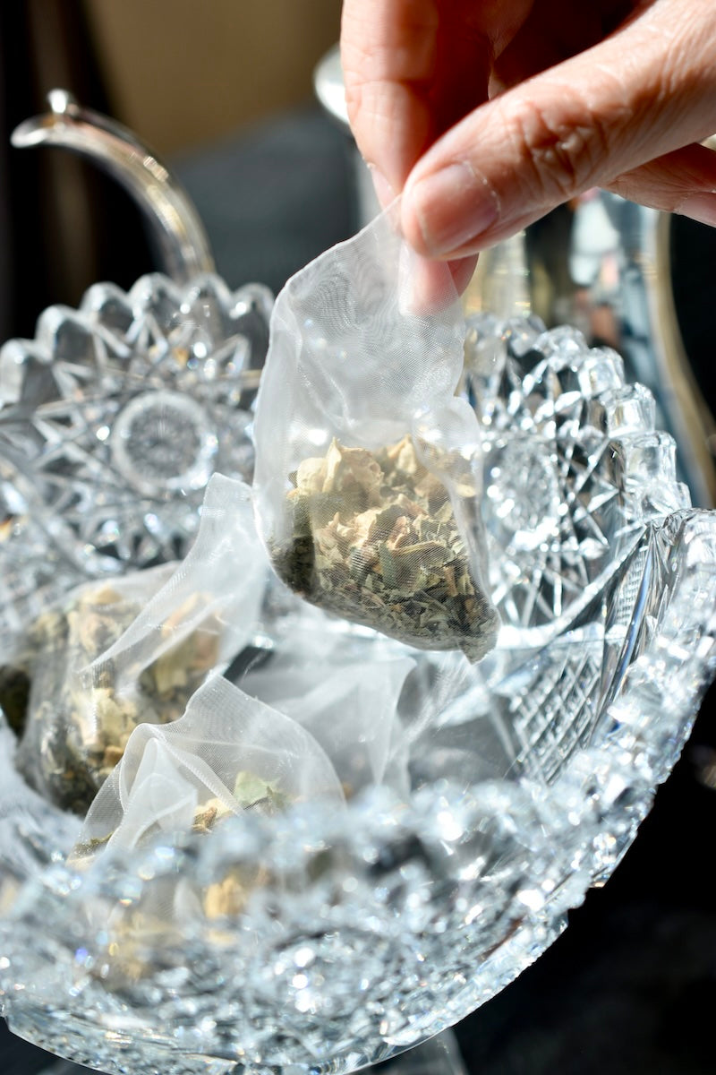 A hand lifting a whole-ingredient herbal sachet from a cut-glass bowl.